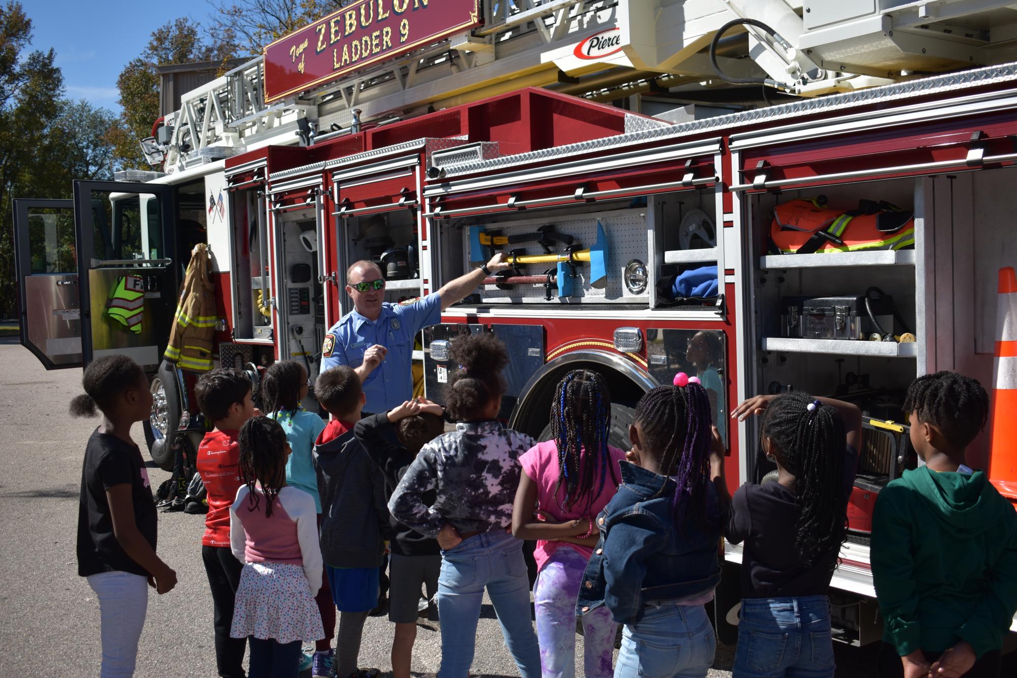 A ZFD firefighter performing an equipment showcase at his fire engine, presenting to a group of local students.